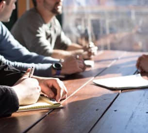 Workers sitting on a large wooden desk having a meeting, each man is taking notes